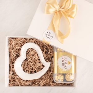 Zestaw prezentowy w białym pudełku z żółtą wstążką, zawierający ceramiczne serce i opakowanie pralinek Ferrero Rocher na tle brązowego papieru wypełniającego.