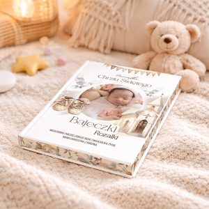 Książka dla dziecka z okładką przedstawiającą śpiące niemowlę w otoczeniu róż, w złotych bucikach, na tle ozdobnej architektury i ptaków, leżąca na miękkim kocu w jasnych kolorach z maskotką misia obok.