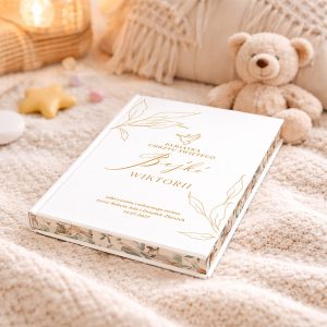 Książka o białej okładce z złotym napisem 'Pamiątka Chrztu Świętego' i imieniem 'Wiktorii', leżąca na jasnym, miękkim kocu obok pluszowego misia i dekoracyjnych elementów takich jak gwiazdka i kolorowe cukierki.