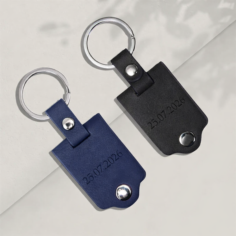 Dwa breloki do kluczy wykonane z materiału syntetycznego, jeden niebieski z grawerem daty 25.07.2026, a drugi czarny z tym samym grawerem, oba z metalowym kółkiem.