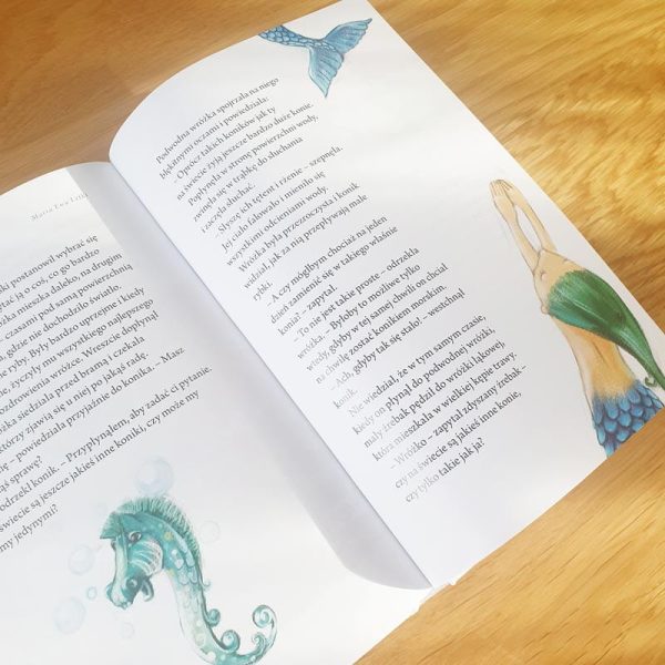 Otwarte strony książki leżące na drewnianym stole, z tekstem opisującym morskie stwory. Na jednej stronie znajduje się ilustracja syreny z zieloną ogonem, na drugiej ilustracja konika morskiego w niebieskich odcieniach, oraz fragment tekstu dotyczący tej tematyki.