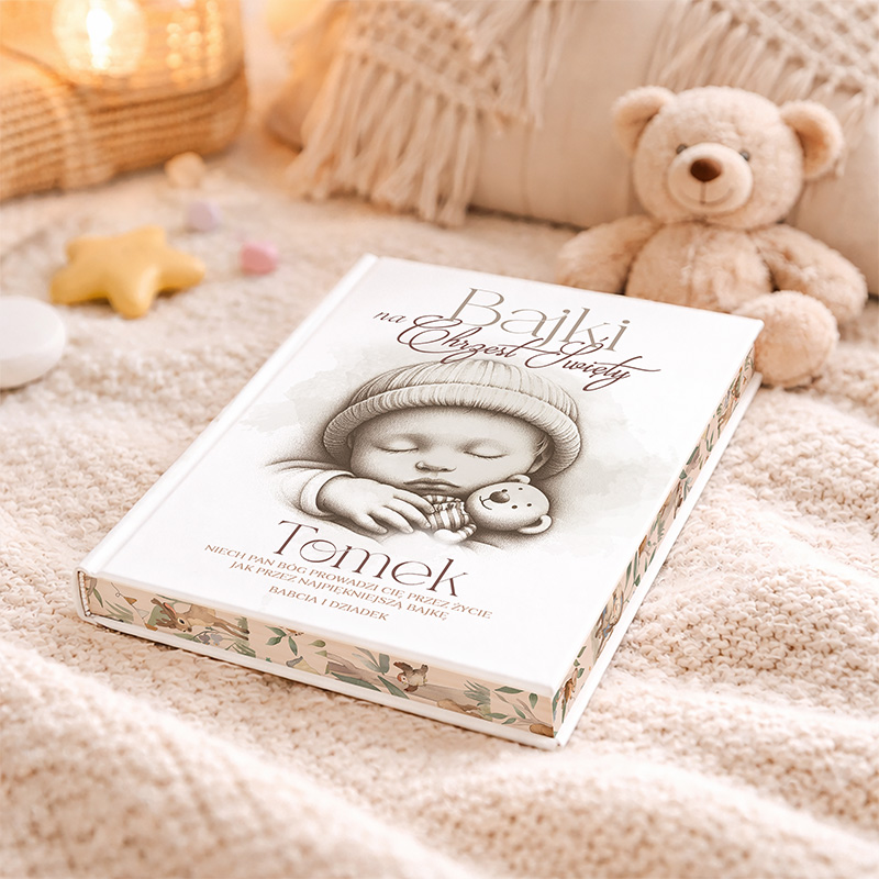 Książka 'Bajki na chrzest święty' leży na jasnym, miękkim kocyku obok pluszowego misia. Okładka książki przedstawia śpiącą postać dziecka z zabawką w ręku, a na niej widnieje imię 'Tomek'. W tle widoczne są dekoracje w formie gwiazdek i lampionów.