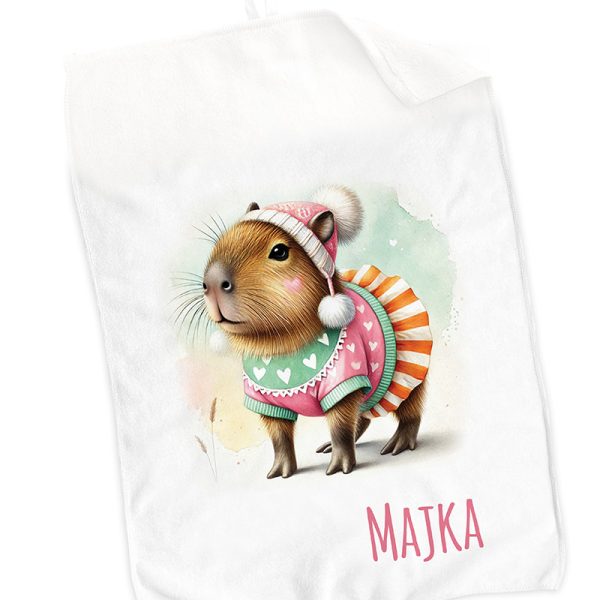 Ilustracja kapibary ubranego w kolorowy sweter z serduszkami i czapkę z pomponem, na tle pastelowych barw, z imieniem "MAJKA" na dole.