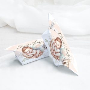Małe pakunki z ilustracją śpiącego niemowlęcia z anielskimi skrzydłami i ręką trzymającą je, na tle jasnego materiału, z napisem 'Chrzest Święty'.