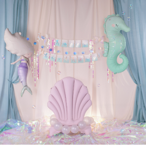 Dekoracja urodzinowa w stylu morskim z motywem syreny oraz konika morskiego, w tle baner "Happy Birthday". W centrum znajduje się różowa, dmuchana muszla jako fotel, otoczona balonami.