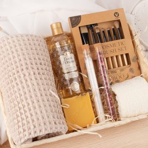 Koszyk z produktami do pielęgnacji zawierający olejek pod prysznic, zestaw pędzli kosmetycznych, dwa pojemniki ze składnikami, mydło, beżowy ręcznik, oraz szary kocyk.