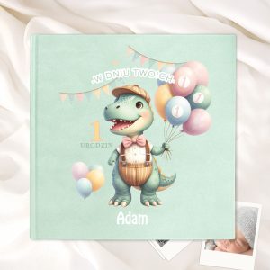 Okładka książki na zielonym tle z ilustracją dinozaura w kapeluszu, trzymającego balony, z napisem 'W Dniu Twoich 1 Urodzin' oraz imieniem 'Adam'.