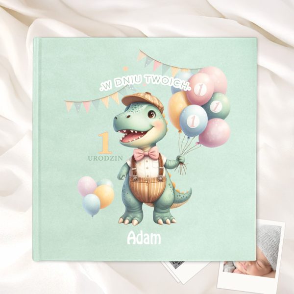 Okładka książki na zielonym tle z ilustracją dinozaura w kapeluszu, trzymającego balony, z napisem 'W Dniu Twoich 1 Urodzin' oraz imieniem 'Adam'.