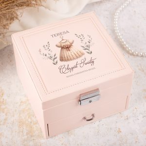 Różowa skrzynka z ozdobnym wieczkiem, na którym znajduje się ilustracja sukienki oraz napisy "TERESA", "Chrzest Święty", i "Mama Chrzestna Beata". Skrzynka ma metalowy zamek i uchwyt z przodu.