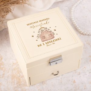 Skrzynia na skarby z napisem "Skrzynia skarbów Marcelinki na 1 urodzinki" oraz wzorem torebki. Elegancka, jasna kolorystyka.