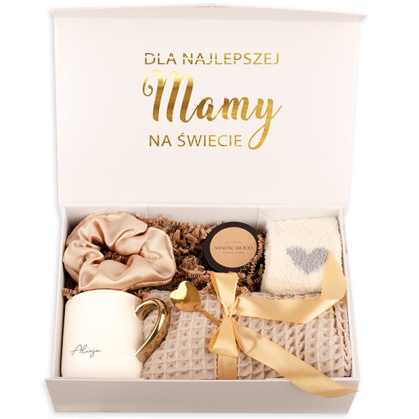 Elegancka biało-złota skrzynka prezentowa z napisem 'DLA NAJLEPSZEJ Mamy NA ŚWIECIE', wypełniona różnymi akcesoriami: satynowym scrunchem w kolorze jasnobeżowym, świecą zapachową o zapachu sandałowym, białym ręcznikiem z szarym sercem, złotą łyżeczką w kształcie serca oraz filiżanką z imieniem 'Aliżja'.