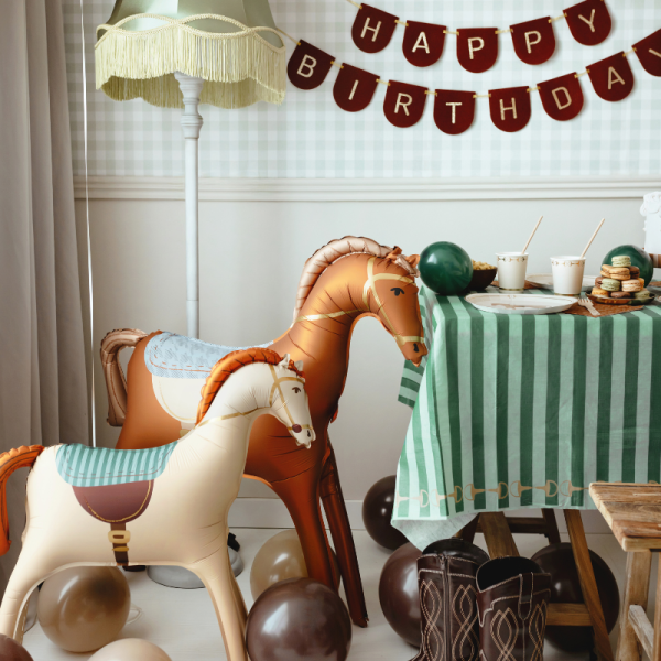 Pokój udekorowany na urodziny, z balonami i oznaczeniem 'HAPPY BIRTHDAY'. W centrum znajdują się dwa nadmuchiwane koniki, a na stole z zielonymi paskami stoją kubki i talerz z ciasteczkami.