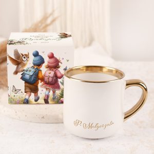 Biała filiżanka z metalowym złotym brzegiem, na której widnieje imię 'P. Małgorzata', obok niej kolorowe opakowanie z ilustracją dzieci w zimowych ubraniach niosących plecaki, otoczonych motylami i sową, na neutralnym tle.