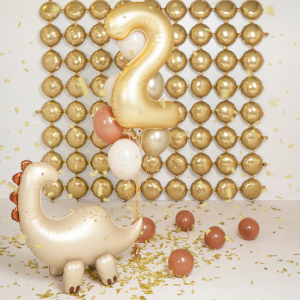 Złoty balon w kształcie cyfry 2 unosi się nad balonową dekoracją w kolorze złotym, z dinozaurem w odcieniach złota i brązu na pierwszym planie, a wokół rozrzucone balony i konfetti na białej podłodze.