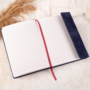 Otwarty notatnik z białymi stronami w kratkę, leżący na jasnym tle, z granatową okładką i czerwoną wstążką jako zakładką, obok trzcinowe dekoracje.