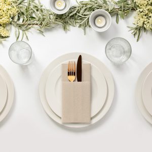 Stół nakryty do posiłku, z trzema talerzami, na każdym z nich elegancko złożony serwetka z widelcem i nożem; w tle dekoracyjne gałęzie roślin oraz świeczniki z podświetleniem.