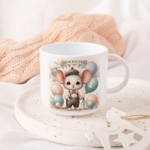 Plastikowy kubek z ilustracją małej myszy w eleganckim ubranku, trzymającej balony, z napisem 'MAM ROCZEK' oraz liczbą '1', umieszczona na talerzu w otoczeniu jasnych materiałów tekstylnych.