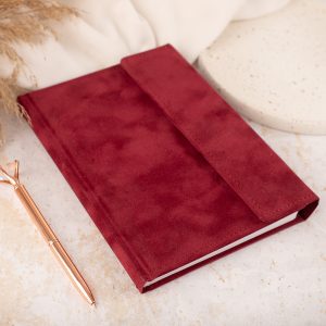 Czerwony notes z okładką z materiału, leżący na powierzchni w kolorze beżowym, obok długopis w kolorze różowego złota oraz neutralne dekoracje na tle.