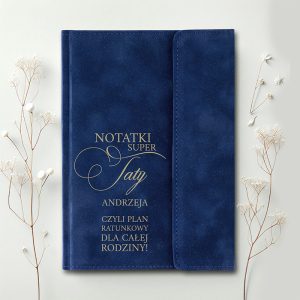Granatowy notes z napisem 'NOTATKI SUPER TATY ANDRZEJA, CZYLI PLAN RATUNKOWY DLA CAŁEJ RODZINY!' w złotym kolorze, umieszczony na jasnym tle obok delikatnych gałązek z kwiatami.