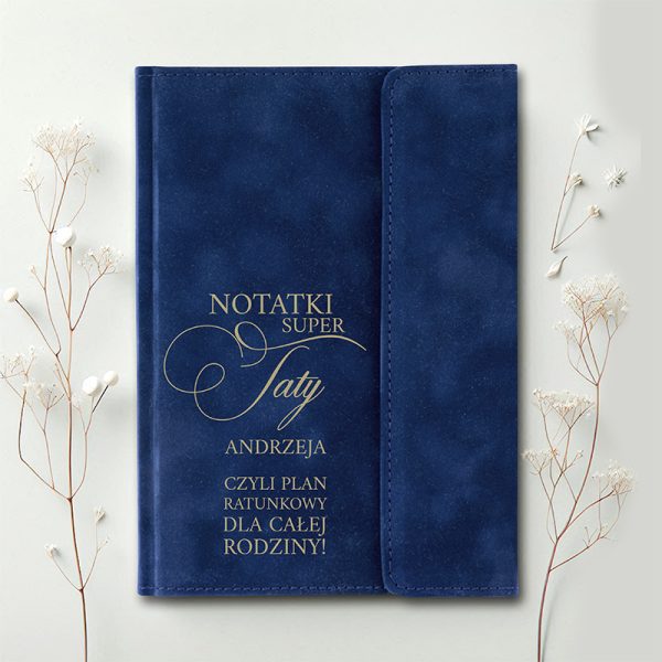 Granatowy notes z napisem 'NOTATKI SUPER TATY ANDRZEJA, CZYLI PLAN RATUNKOWY DLA CAŁEJ RODZINY!' w złotym kolorze, umieszczony na jasnym tle obok delikatnych gałązek z kwiatami.