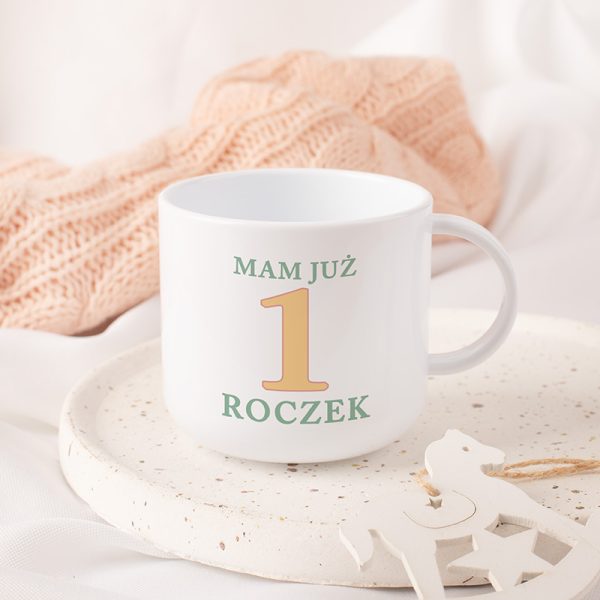 Biały kubek z napisem "Mam już 1 roczek" na tle kremowej talerza i różowego swetra, z białym konikiem na biegunach obok.