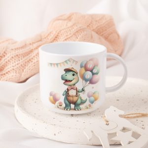 Ceramiczna filiżanka z rysunkiem uśmiechającego się dinozaura w kapeluszu trzymającego kolorowe balony z cyfrą 1, otoczona pastelowym tłem i dekoracjami, na talerzyku z drobnymi czerwonymi i żółtymi wzorami, w otoczeniu miękkich tkanin.