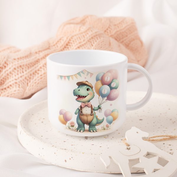 Ceramiczna filiżanka z rysunkiem uśmiechającego się dinozaura w kapeluszu trzymającego kolorowe balony z cyfrą 1, otoczona pastelowym tłem i dekoracjami, na talerzyku z drobnymi czerwonymi i żółtymi wzorami, w otoczeniu miękkich tkanin.