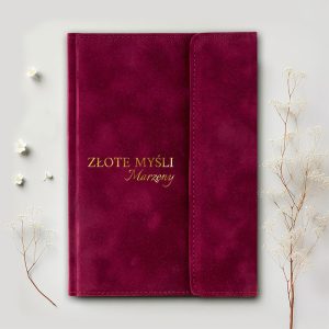 Czerwony notes z tekstem 'ZŁOTE MYŚLI Marzenia' napisanym złotymi literami, leżący na jasnym tle obok białych kwiatków.