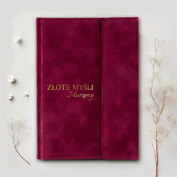Czerwony notes z tekstem 'ZŁOTE MYŚLI Marzenia' napisanym złotymi literami, leżący na jasnym tle obok białych kwiatków.