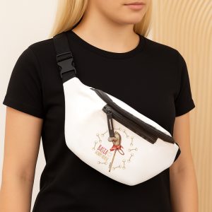 Kobieta z długimi blond włosami, nosząca czarną koszulkę, trzyma na ramieniu biały plecak typu saszetka z grafika przedstawiającą konika hobby i napisem 'BASIA HOBBY HORSE'. Tło jest jasne, z delikatnymi wzorami.