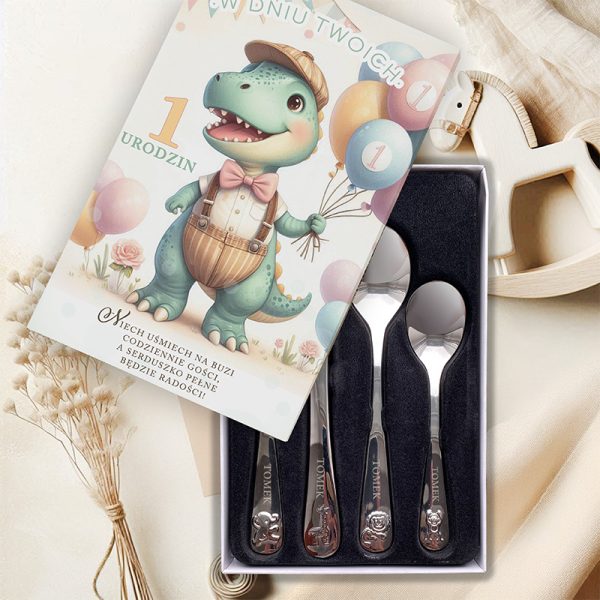 Kartka urodzinowa z ilustracją dinozaura w kapeluszu trzymającego balony, obok zestaw czterech srebrnych łyżeczek w eleganckim pudełku, na tle jasnych tkanin i dekoracji.