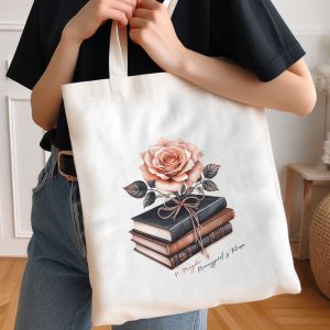 Kobieta trzyma torbę na ramię, na której znajduje się ilustracja różowej róży umieszczonej na stercie książek. Torba jest jasna, a w tle widać fragment wnętrza mieszkania.