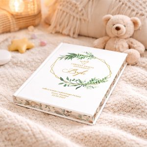 Ozdobna książka z białą okładką i złotym napisem, leżąca na jasnym kocyku obok pluszowego misia; w tle widać delikatne dekoracje i akcesoria. Na okładce znajdują się zielone liście i napis "CHRZEST ŚWIĘTY ANTONIEGO".