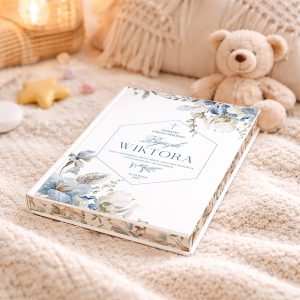 bajki-z-okladka-niebieskich-kwiatow-na-chrzest Książka z okładką w białym kolorze, ozdobioną niebieskimi i zielonymi kwiatami oraz napisem 'Pamiątka Chrztu Świętego Wiktora', leży na jasnym kocie obok pluszowego misia oraz dekoracyjnych przedmiotów, jak gwiazda i małe cukierki.