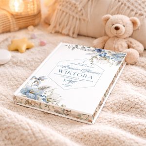 Biała książka z delikatnym wzorem kwiatowym, nosząca napis "WIKTORA" oraz datę 10 sierpnia 2022, leży na jasnym, miękkim kocu obok pluszowego misia i dekoracyjnych elementów, takich jak gwiazda i kamyczki.