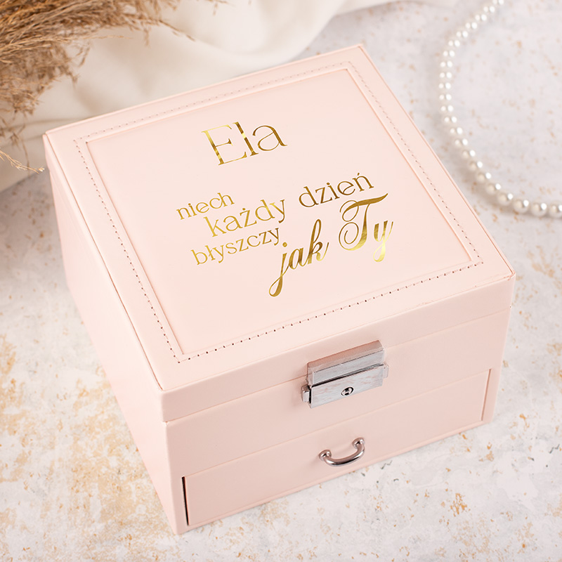 Różowa szkatułka z metalowym zamkiem, na wieczku złote napisy: "Ela niech każdy dzień błyszczy jak Ty". W tle widać delikatne dekoracje, jak perły i suszone kwiaty.