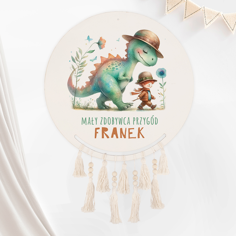 Ilustracja przedstawiająca zielonego dinozaura w kapeluszu i małego chłopca, również w kapeluszu, spacerujących razem wśród kwiatów; widnieje napis "MAŁY ZDOBYWCA PRZYGÓD FRANEK" na okrągłej tablicy z makramową ozdobą u dołu.