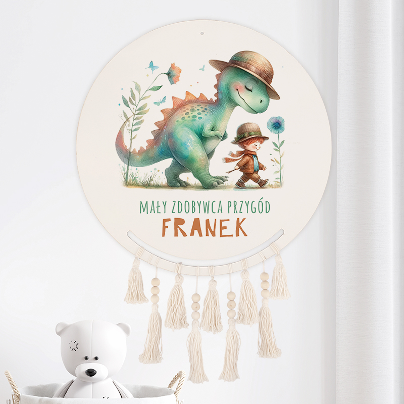 Ilustracja przedstawiająca małego dinozaura w kapeluszu, idącego z chłopcem, również w kapeluszu, pośród kwiatów. W górnej części napisy "MAŁY ZDOBYWCA PRZYGÓD" oraz imię "FRANEK". Elementy zdobnicze u dołu w postaci sznurka z frędzlami. Tło jasne, z białymi zasłonami w tle oraz białym misiem zabawką na pierwszym planie.