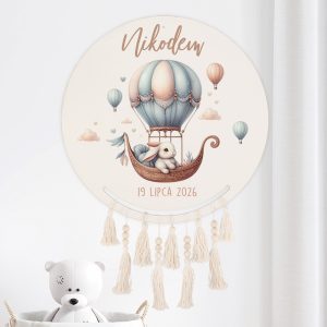 Dekoracyjny okrągły obrazek przedstawiający króliczka w balonie na gorące powietrze, z napisem 'Nikodem' i datą '19 lipca 2026'. Grafika w pastelowych kolorach z dodatkowymi balonami i chmurkami, z frędzlami na dole obrazu. W tle widoczny biały przód zabawki misia.