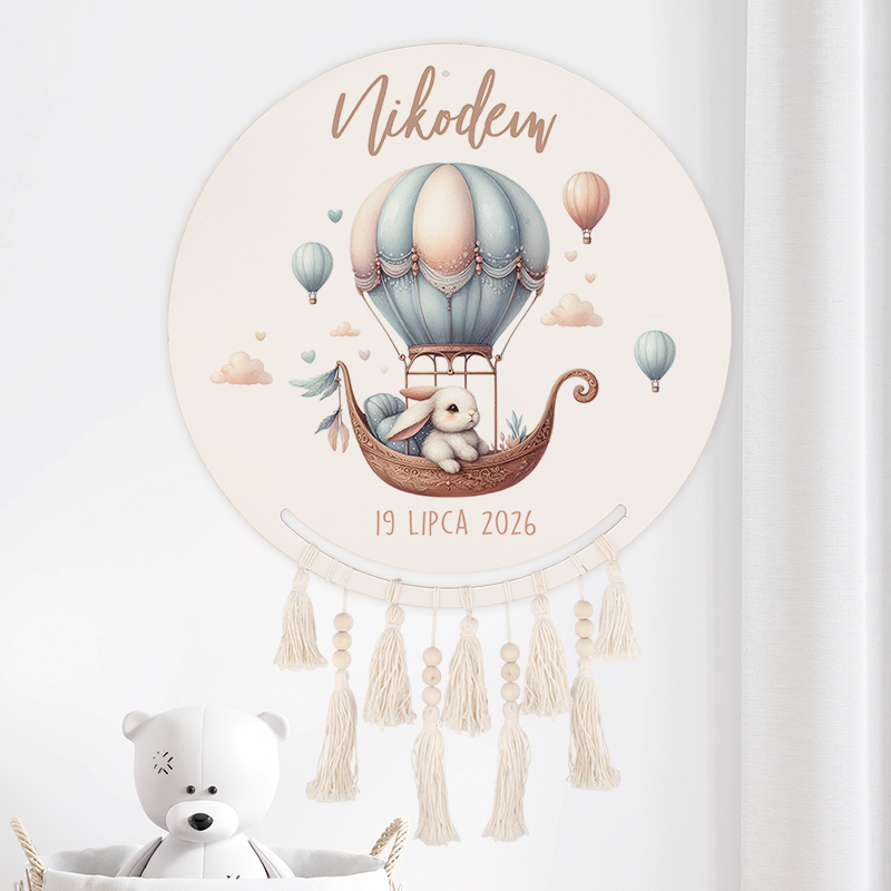 Dekoracyjny okrągły obrazek przedstawiający króliczka w balonie na gorące powietrze, z napisem 'Nikodem' i datą '19 lipca 2026'. Grafika w pastelowych kolorach z dodatkowymi balonami i chmurkami, z frędzlami na dole obrazu. W tle widoczny biały przód zabawki misia.