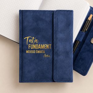 Granatowy notes z napisem "Tata FUNDAMENT mojego świata" oraz imieniem "Ania". Na tle widać pustą kratkowaną stronę i długopis.