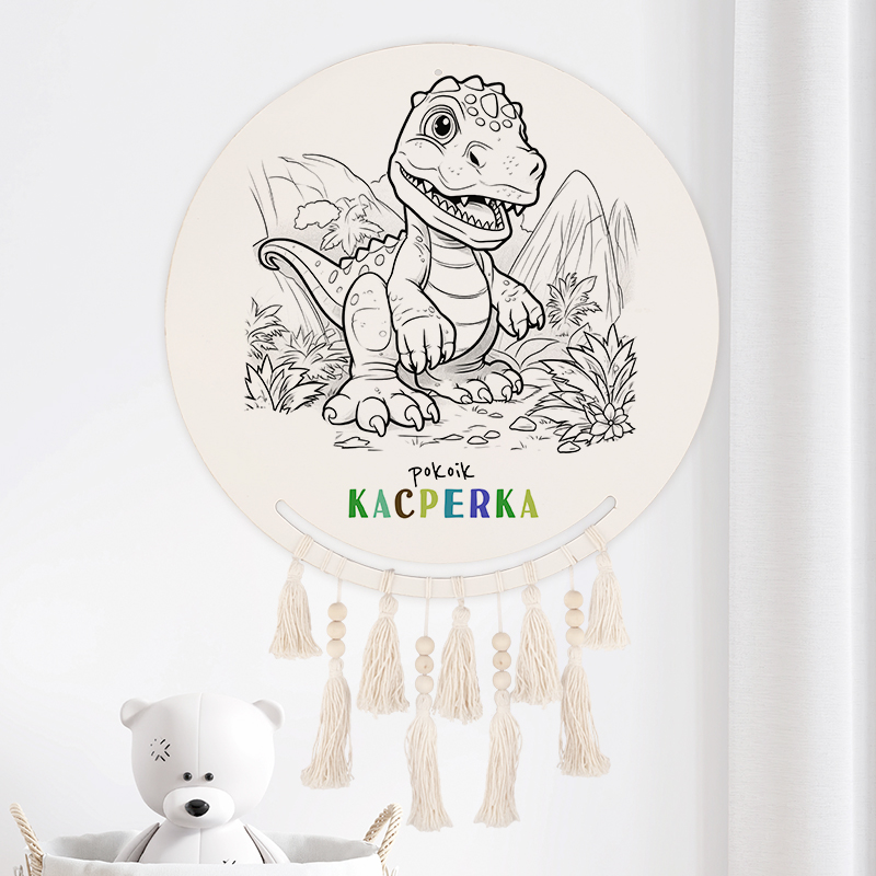Okrągła dekoracja ścienna z konturami dinozaura, otoczona roślinnością, z napisem 'pokoik Kacperka' w kolorze zielonym, żółtym i niebieskim; z frędzlami na dole dekoracji. W tle widoczna pluszowa zabawka w kształcie misia.