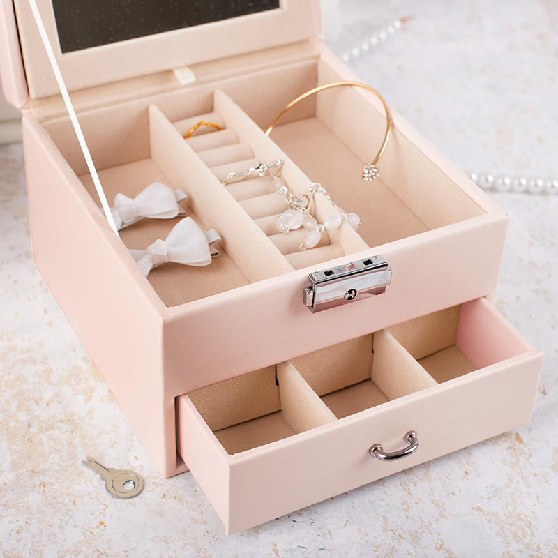 Różowe etui na biżuterię z lustrem, otwarte, z kolczykami, pierścionkami i białymi kokardkami w środku, a także z wysuwanym szufladką i kluczykiem obok.