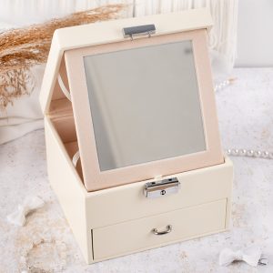 szkatulka-na-bizuterie-lusterko-kremowa Elegancka kremowa szkatułka na biżuterię z lustrem i szufladką, na tle jasnego materiału i biżuterii perłowej.