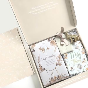 Otwarta pudełko z dekoracyjnymi przedmiotami, w tym zaproszeniem do chrztu, książką zatytułowaną 'Mój Pierwszy Spacer', małym, białym woreczkiem ozdobionym kokardką oraz metalowym słoniem, na delikatnym beżowym tle z wytłoczonymi liśćmi.