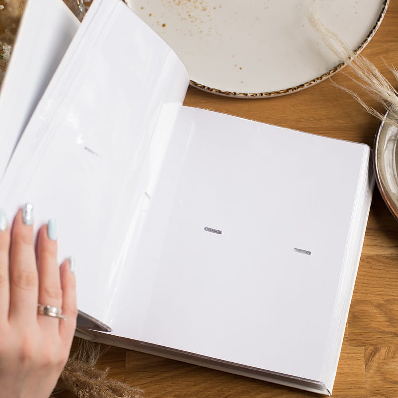 Widok na otwartą, pustą teczkę z przezroczystymi kieszonkami, na której znajduje się dłoń z pomalowanymi na jasno niebiesko paznokciami. W tle widać talerz z brzegiem w brązowym kolorze oraz fragmenty roślinnych ozdób na drewnianym blacie.