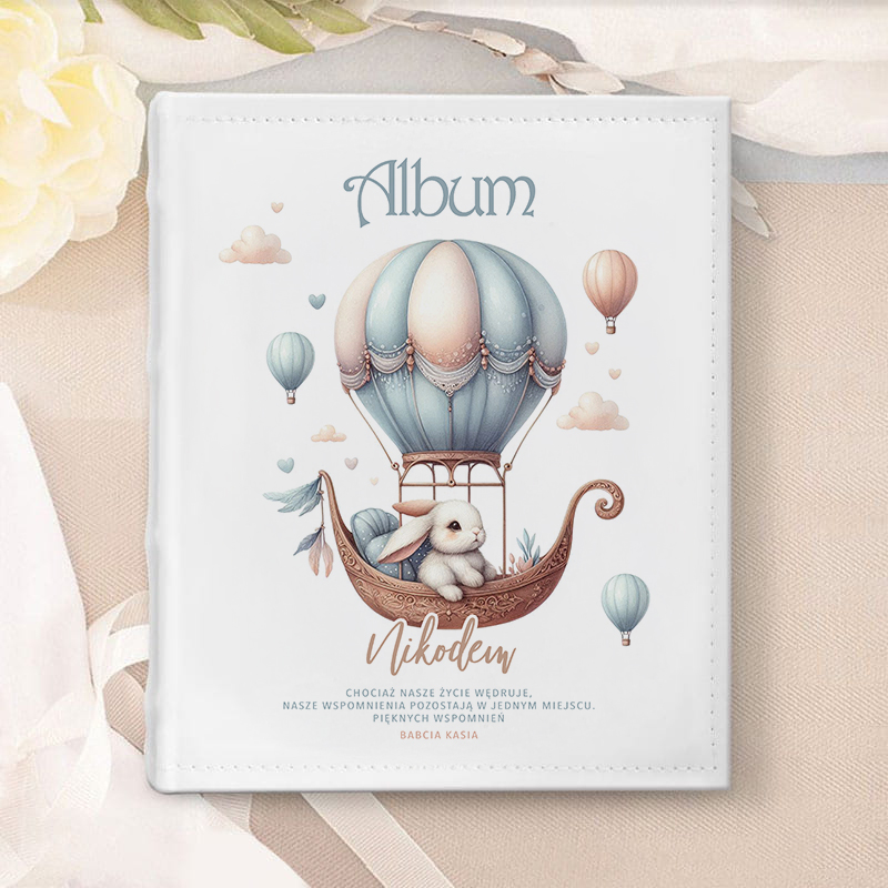 Książka na wspomnienia z białą okładką, na której znajduje się ilustracja balonu na gorące powietrze z króliczkiem siedzącym w koszyku. W tle unoszą się kolorowe baloniki oraz chmurki. Na okładce napis "Album" i imię "Nikodem", poniżej tekst w języku polskim.