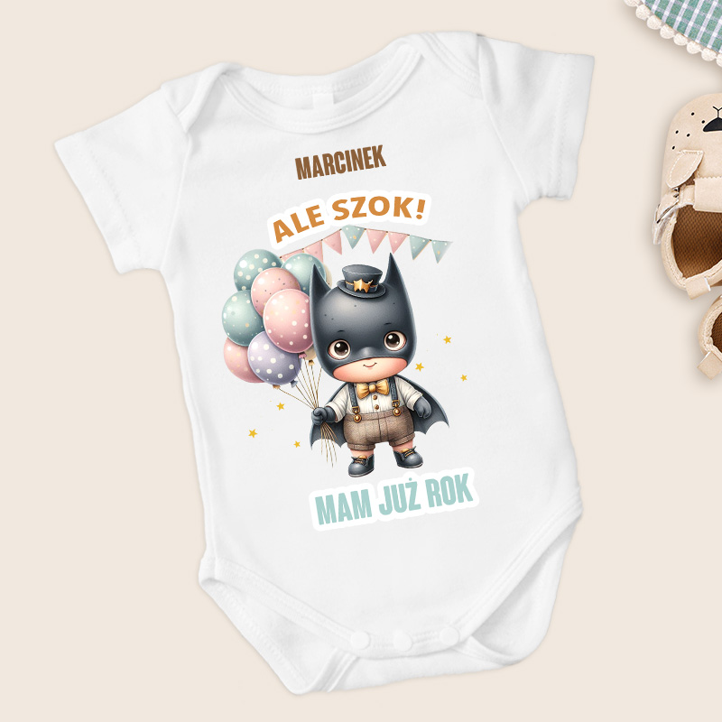 Białe body dla dzieci z ilustracją uroczego chłopca w przebraniu superbohatera, trzymającego balony w różnych kolorach, z napisem "Marcinek ALE SZOK! MAM JUŻ ROK" na górze i dole.
