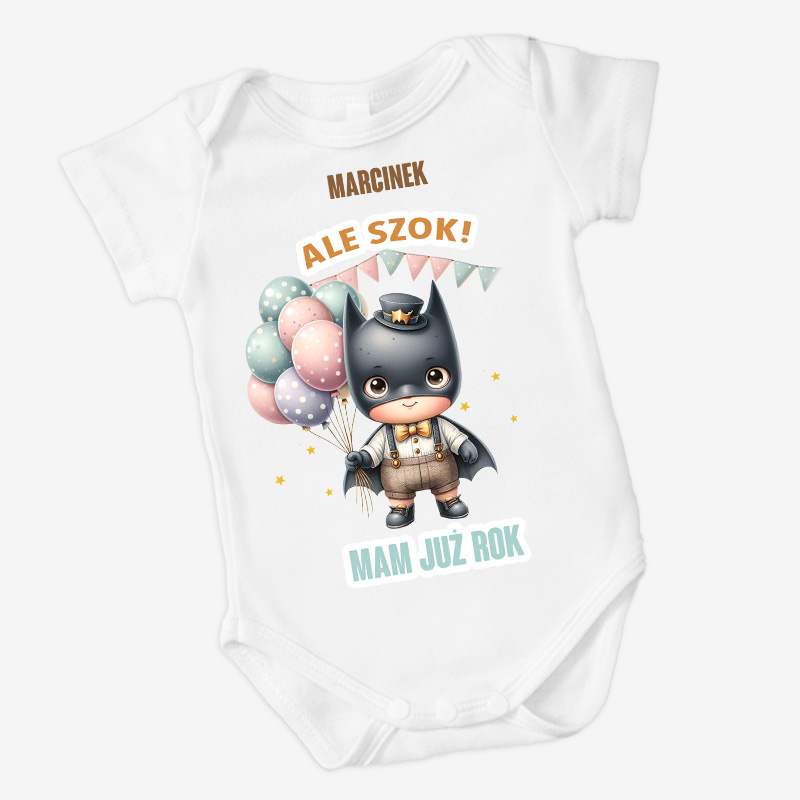 Biały body dziecięce z ilustracją słodkiego, małego bohatera w kostiumie nietoperza, trzymającego balony w pastelowych kolorach. Na górze napis 'MARCINEK' oraz 'ALE SZOK!', a na dole 'MAM JUŻ ROK'.