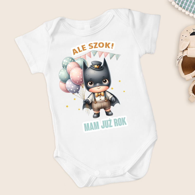 Biały body dla dzieci z ilustracją małego superbohatera w czarnym kostiumie oraz kapeluszu, trzymającego kolorowe balony. Na koszulce napis: 'ALE SZOK!' i 'MAM JUŻ ROK'.